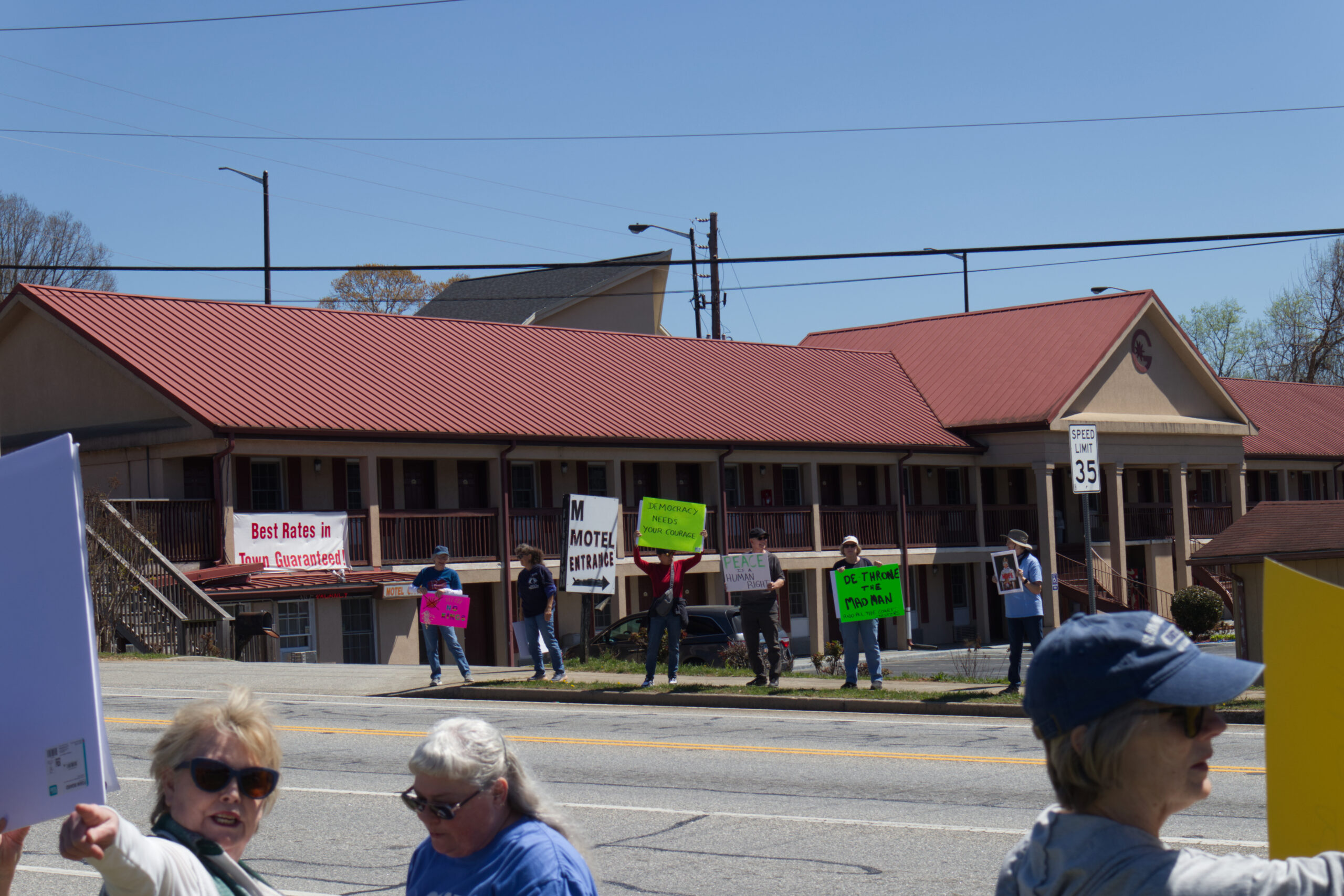 Protesters stand in front of a motel holding signs that read. From left to right: "No Kings", "Democracy Needs your courage","Peace is a human right", De-Throne the Mad Man"